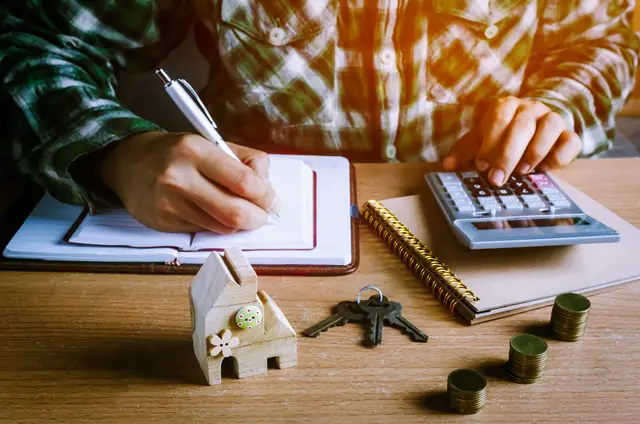 Pessoa fazendo cálculos ao lado de uma miniatura de casa e moedas